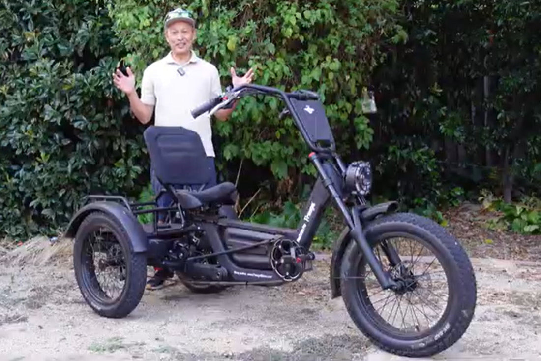 Side profile of the FlekTrike Pro All-Terrain Tilting eTrike with backrest, showing tires, handlebars, headlight, front fender, rear dual wheels,and comfortable seated position, with a smiling rider posing beside it in a green garden/outdoor setting.
