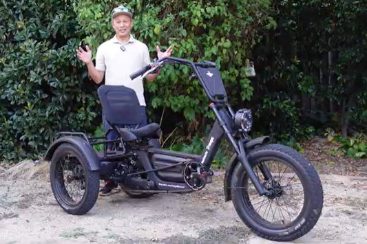 Side profile of the FlekTrike Pro All-Terrain Tilting eTrike with backrest, showing tires, handlebars, headlight, front fender, rear dual wheels,and comfortable seated position, with a smiling rider posing beside it in a green garden/outdoor setting.