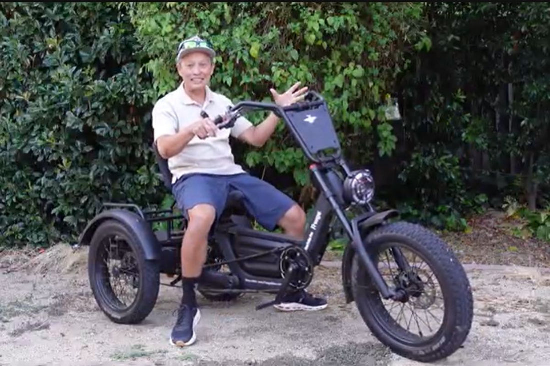 Smiling senior man sitting on a black Fat Tire electric trike with a front storage basket, parked outdoors against green bushes, demonstrating convenience for electric trike storage and charging.