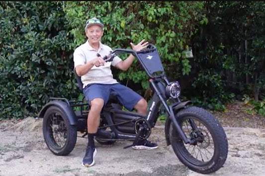 Smiling senior man sitting on a black Fat Tire electric trike with a front storage basket, parked outdoors against green bushes, demonstrating convenience for electric trike storage and charging.
