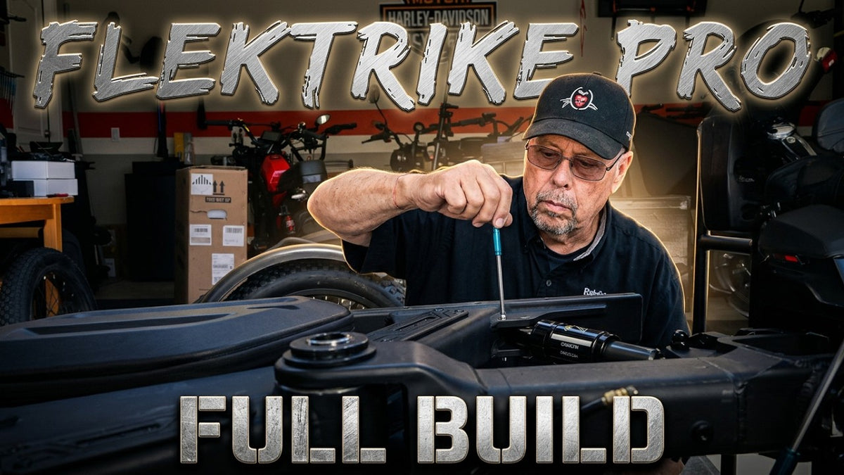 A man in a garage wearing a black cap and dark shirt works on FlekTrike Pro All-Terrain Tilting eTrike, with large metallic text reading 'FLEKTRIKE PRO' at the top and 'FULL BUILD' at the bottom.