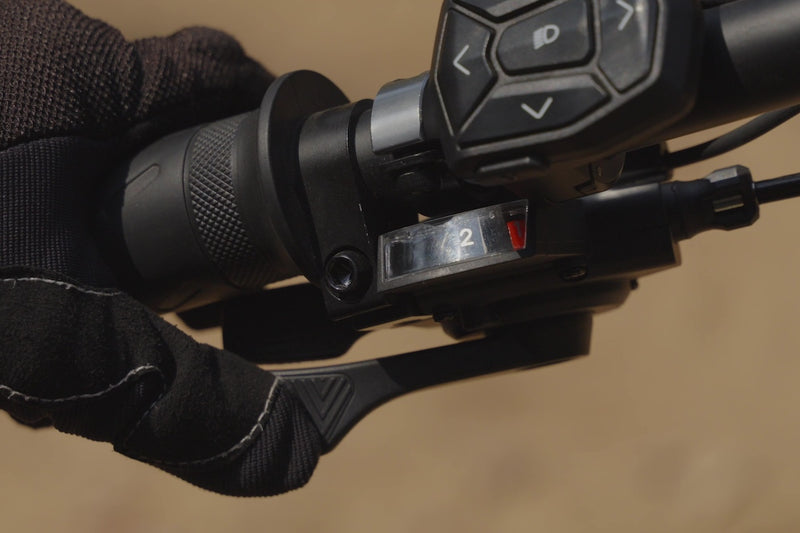 Close-up of a black bicycle handlebar control module with a gloved hand holding the lever, set against a blurred tan background.